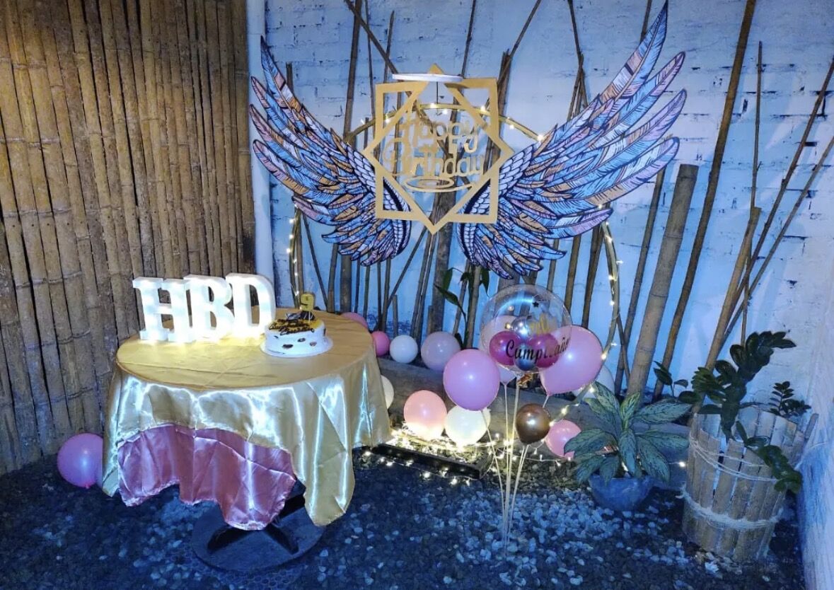 Mesa de cumpleaños con alas decorativas, globos rosados y letras HBD iluminadas